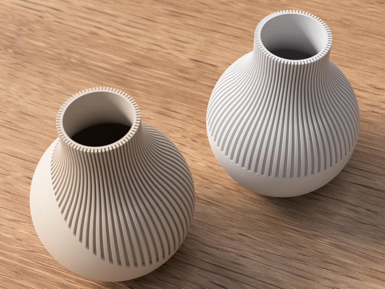 May include: Two off-white, modern vases with a ribbed design. The vases have a rounded base and a cylindrical opening. The ribbed pattern covers the body of the vases, creating a textured look. They sit on a light brown wooden surface.