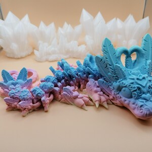 May include: A 3D-printed dragon figurine in shades of blue, pink, and purple. The dragon has intricate details, including feathered wings and a detailed head. White crystal formations are in the background. The dragon is a fantasy-themed item.