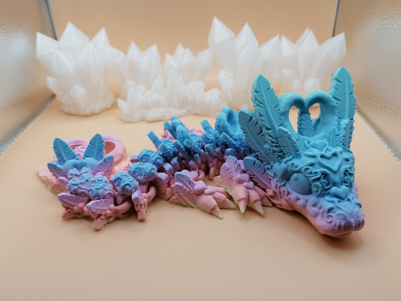 May include: A 3D-printed dragon figurine in shades of blue, pink, and purple. The dragon has intricate details, including feathered wings and a detailed head. White crystal formations are in the background. The dragon is a fantasy-themed item.