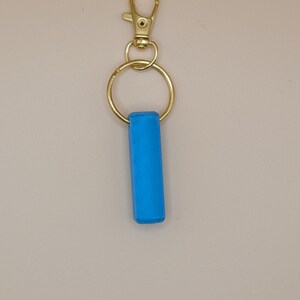 May include: A bright blue rectangular keychain charm attached to a gold-colored metal keyring and clasp. The charm is a solid, vibrant blue. The clasp is a lobster claw style.
