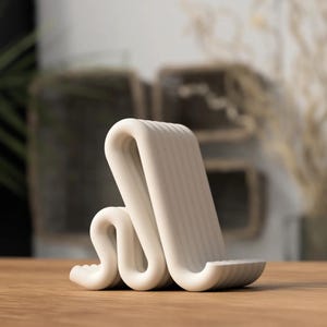 Puede incluir: Un soporte para tel&eacute;fono blanco y moderno con un dise&ntilde;o ondulado &uacute;nico. El soporte est&aacute; hecho de un material liso y s&oacute;lido y presenta una textura acanalada en el soporte en &aacute;ngulo y la base. Se encuentra sobre una superficie de madera.