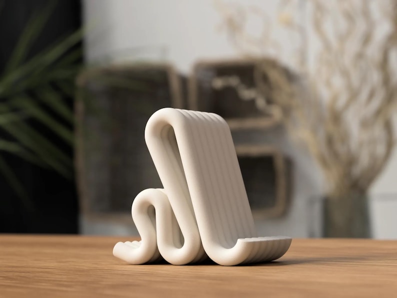 Puede incluir: Un soporte para tel&eacute;fono blanco y moderno con un dise&ntilde;o ondulado &uacute;nico. El soporte est&aacute; hecho de un material liso y s&oacute;lido y presenta una textura acanalada en el soporte en &aacute;ngulo y la base. Se encuentra sobre una superficie de madera.