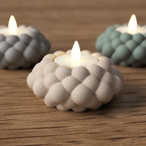 May include: Three decorative candle holders with a textured, bubble-like design. The candle holders are in shades of gray, white, and teal, each holding a lit, battery-operated candle. The holders sit on a wooden surface.