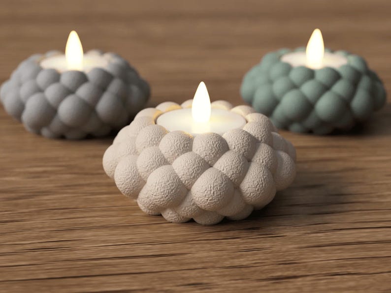 May include: Three decorative candle holders with a textured, bubble-like design. The candle holders are in shades of gray, white, and teal, each holding a lit, battery-operated candle. The holders sit on a wooden surface.