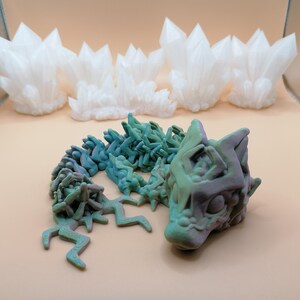 May include: A 3D-printed articulated dragon in shades of green and purple, with a detailed head and body. Behind it are several white, crystalline formations, resembling clusters of crystals. The objects are set against a neutral background.