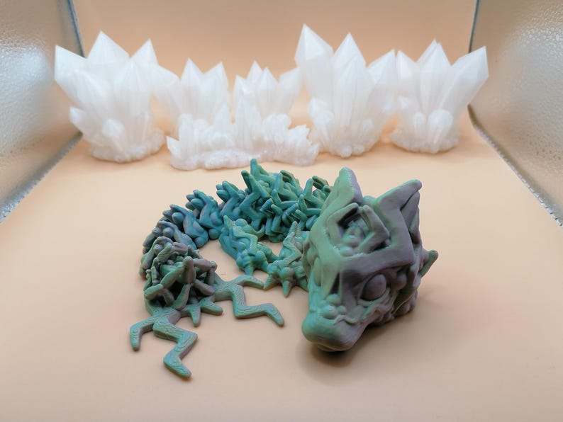 May include: A 3D-printed articulated dragon in shades of green and purple, with a detailed head and body. Behind it are several white, crystalline formations, resembling clusters of crystals. The objects are set against a neutral background.