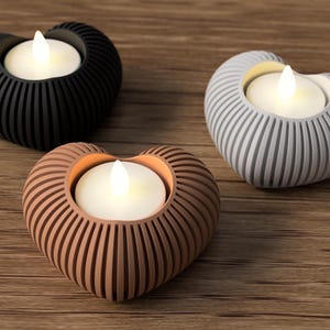 May include: Three heart-shaped candle holders in black, gray, and brown, each with a white LED candle. The holders have a ribbed design and sit on a wooden surface. A decorative accent for home decor.
