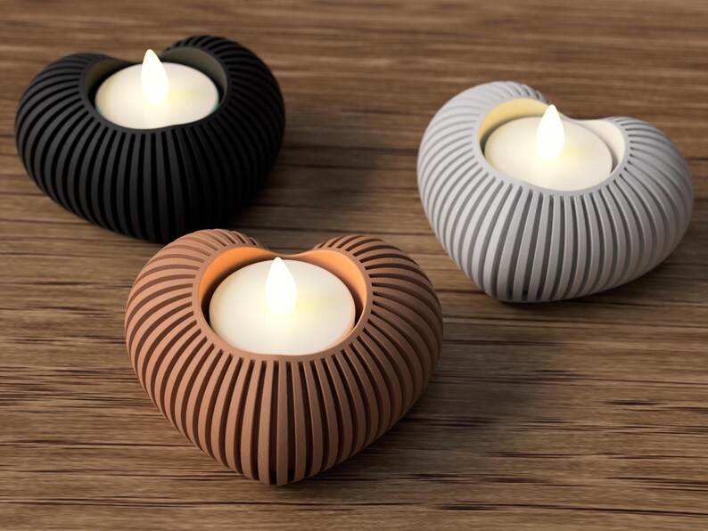 May include: Three heart-shaped candle holders in black, gray, and brown, each with a white LED candle. The holders have a ribbed design and sit on a wooden surface. A decorative accent for home decor.
