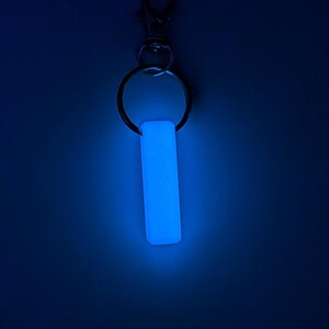 May include: A glowing blue rectangular keychain charm. The charm is attached to a silver-colored keyring and a lobster clasp. The background is a dark blue.