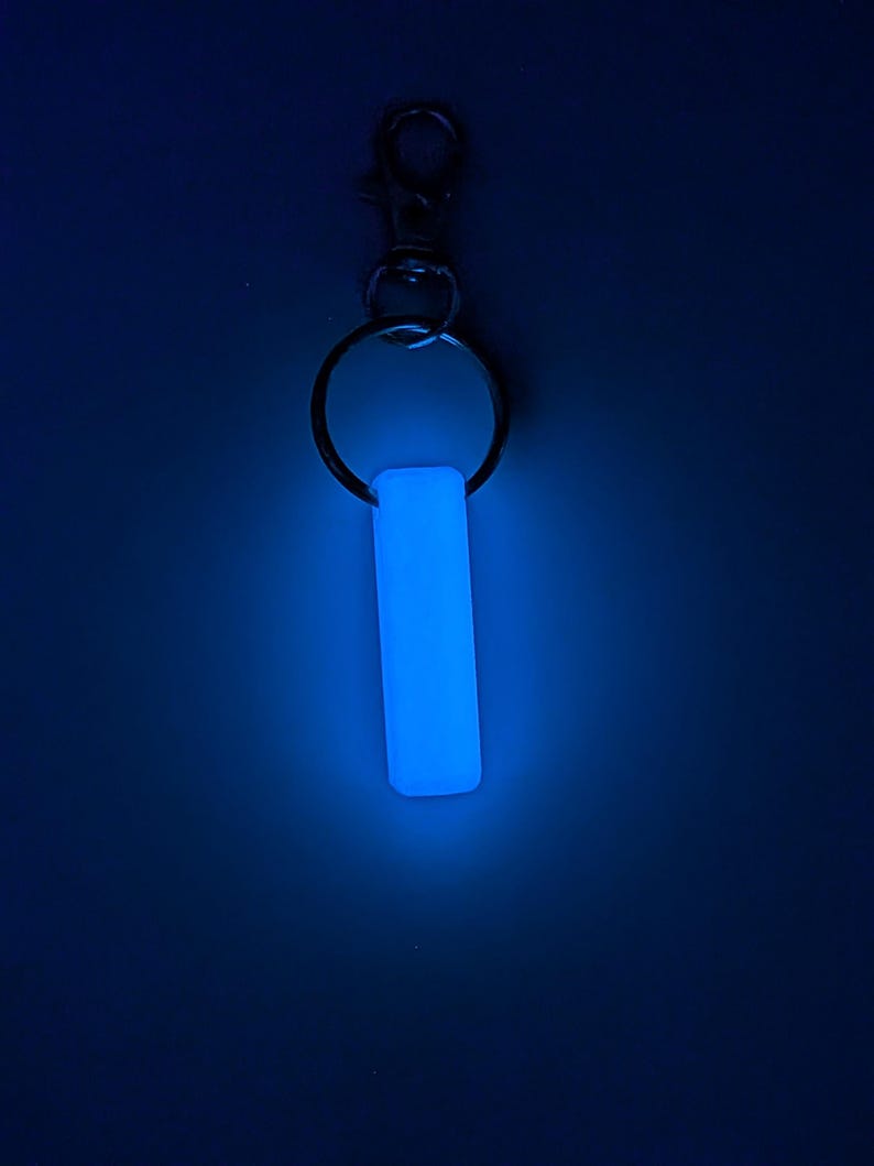 May include: A glowing blue rectangular keychain charm. The charm is attached to a silver-colored keyring and a lobster clasp. The background is a dark blue.