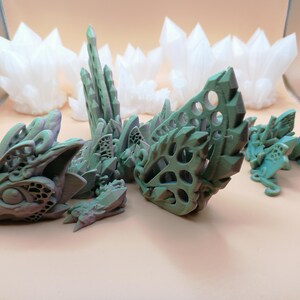 Pu&ograve; includere: Una collezione di sculture di draghi fantasy stampate in 3D in tonalit&agrave; di verde e grigio, con intricati disegni traforati. Diverse strutture bianche a forma di cristallo sono sullo sfondo, creando un tema fantasy.