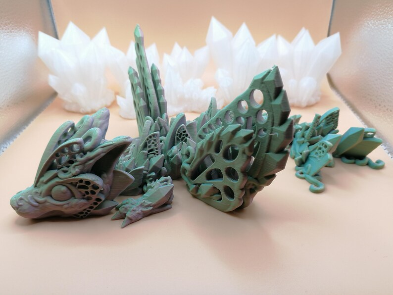 Pu&ograve; includere: Una collezione di sculture di draghi fantasy stampate in 3D in tonalit&agrave; di verde e grigio, con intricati disegni traforati. Diverse strutture bianche a forma di cristallo sono sullo sfondo, creando un tema fantasy.