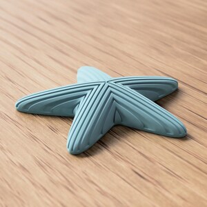 May include: A light blue starfish-shaped object with ribbed detailing. The five-pointed design is set against a wooden surface. The object appears to be made of a solid material.