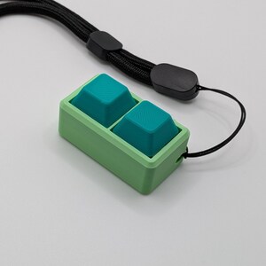 May include: A small, rectangular device with two teal buttons set in a light green frame. A black lanyard is attached to the device. The buttons have a textured surface, and the frame has a smooth finish.