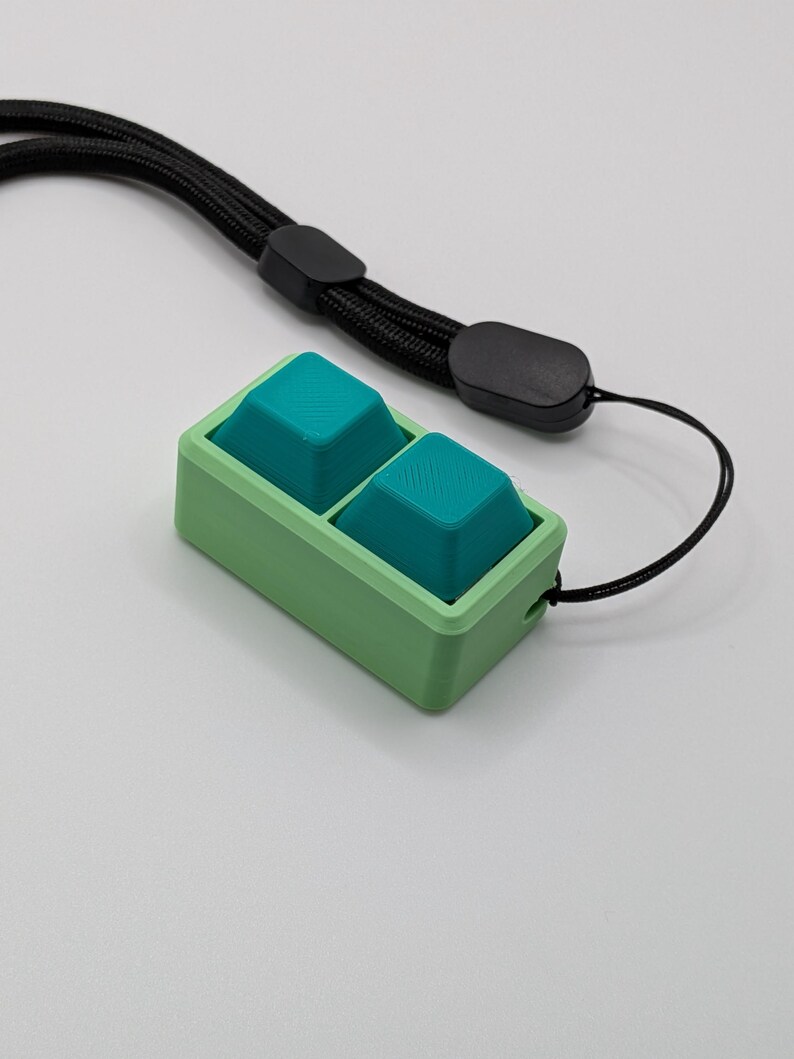 May include: A small, rectangular device with two teal buttons set in a light green frame. A black lanyard is attached to the device. The buttons have a textured surface, and the frame has a smooth finish.