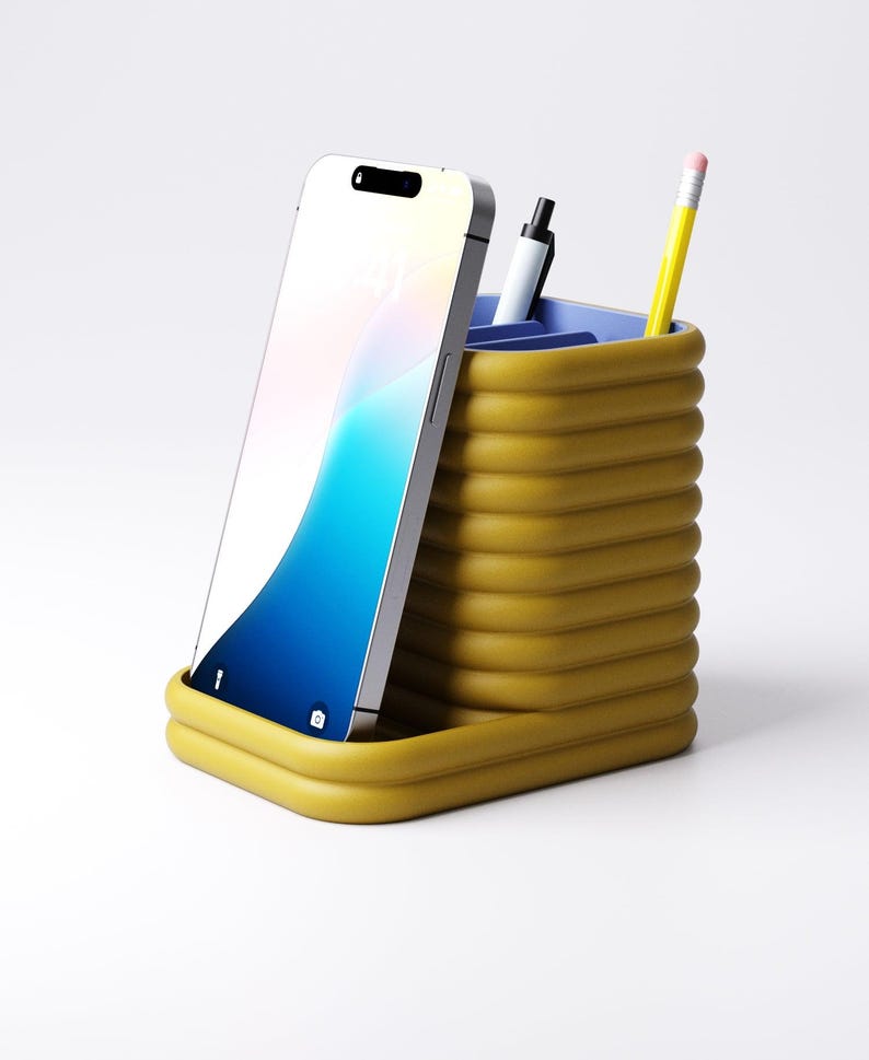 May include: A mustard yellow desk organizer with a ribbed design, holding a smartphone, a black pen, and a yellow pencil with a pink eraser. The organizer has a blue interior and sits on a white surface.