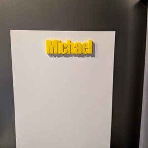 May include: A yellow 3D-printed name, "Michael", is affixed to a white surface. The name is in a bold, sans-serif font. The white surface is mounted on a metallic gray background.