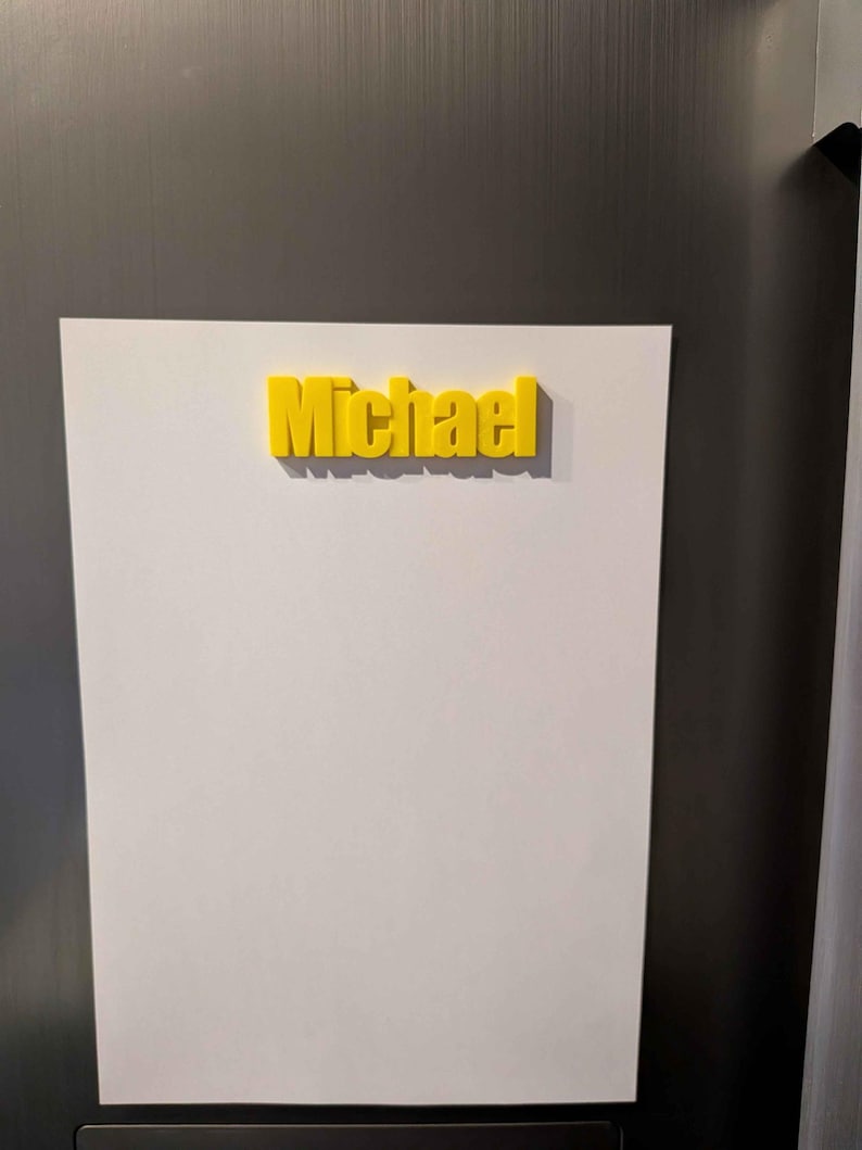 May include: A yellow 3D-printed name, "Michael", is affixed to a white surface. The name is in a bold, sans-serif font. The white surface is mounted on a metallic gray background.