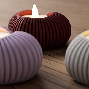 May include: Three decorative candle holders in shades of purple, burgundy, and gray, each with a glowing LED candle. The holders have a ribbed, rounded design. The candles are on a wooden surface.