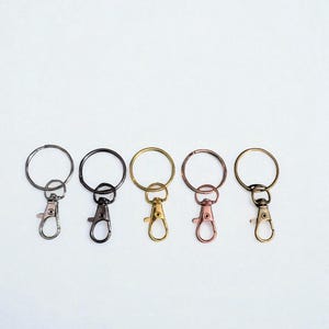 May include: Five keychains with different metal finishes: silver, black, gold, rose gold, and brass. Each keychain has a round ring and a lobster clasp for attaching keys or other items. The keychains are arranged in a row against a white background.