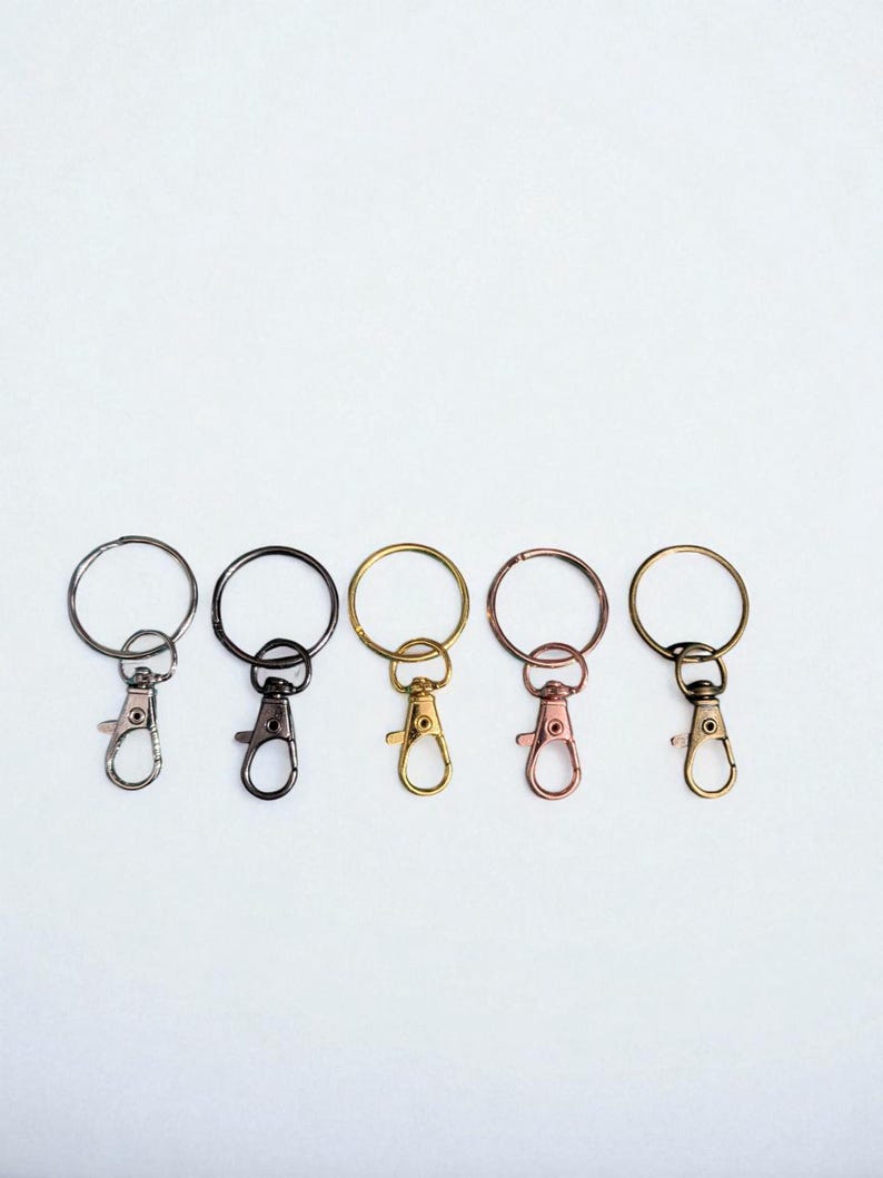 May include: Five keychains with different metal finishes: silver, black, gold, rose gold, and brass. Each keychain has a round ring and a lobster clasp for attaching keys or other items. The keychains are arranged in a row against a white background.