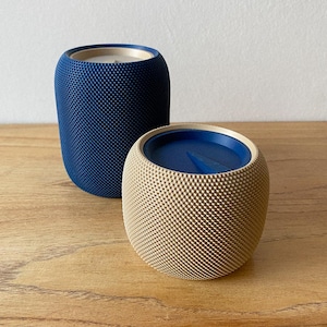 May include: Two cylindrical containers with a textured, dimpled exterior surface. One container is dark blue with a light-coloured interior rim, while the other is beige with a dark blue lid featuring a small triangular indentation on the top surface.