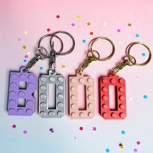 May include: Four keychains spelling out "BOO" in a block-style font. The letters are purple, silver, light pink, and red. Each keychain has a metal ring and clasp. The background is a gradient of pink and blue, with scattered confetti.