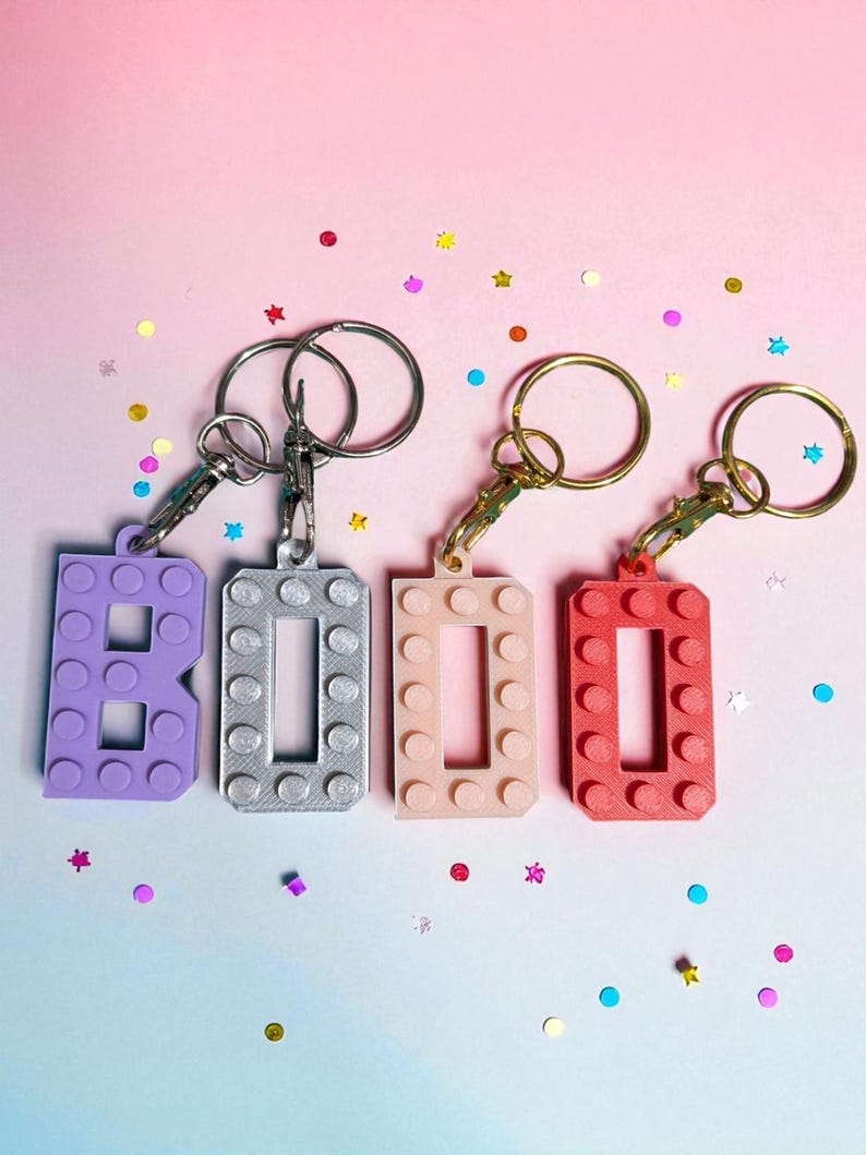 May include: Four keychains spelling out "BOO" in a block-style font. The letters are purple, silver, light pink, and red. Each keychain has a metal ring and clasp. The background is a gradient of pink and blue, with scattered confetti.