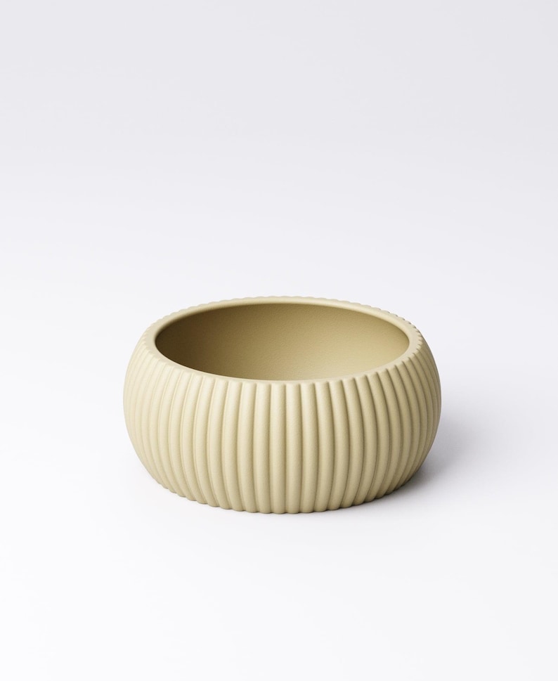 May include: A round, light beige ceramic bowl with vertical ribbed detailing. The bowl has a wide, open interior and a smooth, matte finish. The design is simple and modern, suitable for various uses.