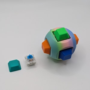 May include: A colorful, spherical puzzle with six cube-shaped pieces in green, blue, and orange. A single teal keycap and a mechanical keyboard switch are also present. The puzzle has a rainbow gradient and is likely 3D-printed.