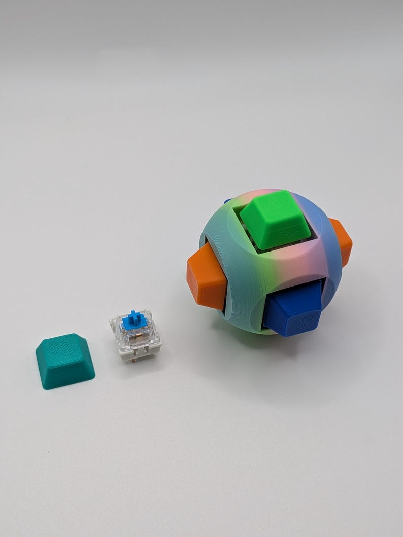 May include: A colorful, spherical puzzle with six cube-shaped pieces in green, blue, and orange. A single teal keycap and a mechanical keyboard switch are also present. The puzzle has a rainbow gradient and is likely 3D-printed.