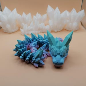 May include: A collection of 3D-printed crystal formations and a dragon figurine. The dragon is blue and purple, with a detailed, geometric design. Several white crystal clusters are in the background, all on a light brown surface.
