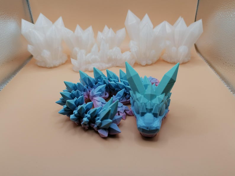 May include: A collection of 3D-printed crystal formations and a dragon figurine. The dragon is blue and purple, with a detailed, geometric design. Several white crystal clusters are in the background, all on a light brown surface.