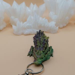 May include: A green, purple, and gold dragon head keychain with a silver keyring. The dragon is 3D printed and has a detailed, textured surface. White, crystalline formations are in the background.
