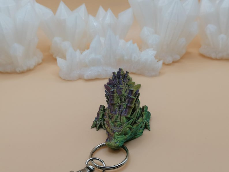 May include: A green, purple, and gold dragon head keychain with a silver keyring. The dragon is 3D printed and has a detailed, textured surface. White, crystalline formations are in the background.