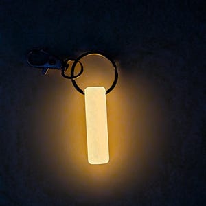 May include: A glowing, rectangular keychain tag with a metal clasp and ring. The tag emits a soft, warm light against a dark background. The keychain is designed for visibility in low-light conditions.