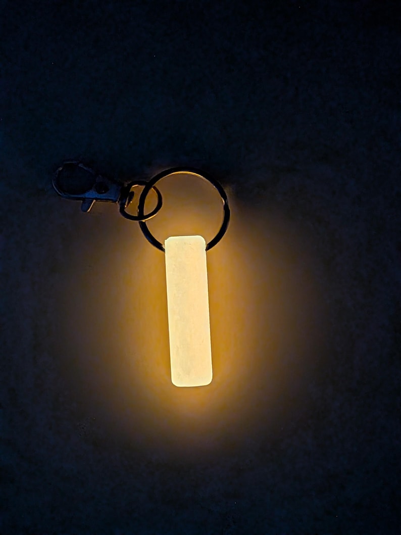 May include: A glowing, rectangular keychain tag with a metal clasp and ring. The tag emits a soft, warm light against a dark background. The keychain is designed for visibility in low-light conditions.