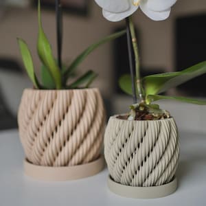 May include: Two round plant pots with a textured, spiralling rope-like pattern on the exterior. Each pot sits on a matching circular saucer. One pot is a light beige colour, and the other is a slightly darker tan, each containing a green plant.