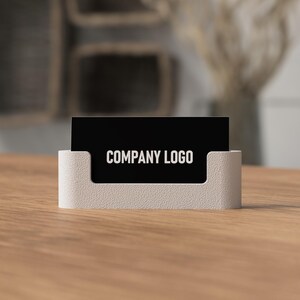 May include: A business card holder with a black card displaying "COMPANY LOGO" in white text. The holder is a light beige color and sits on a wooden surface. The background is blurred, suggesting an office or workspace setting.