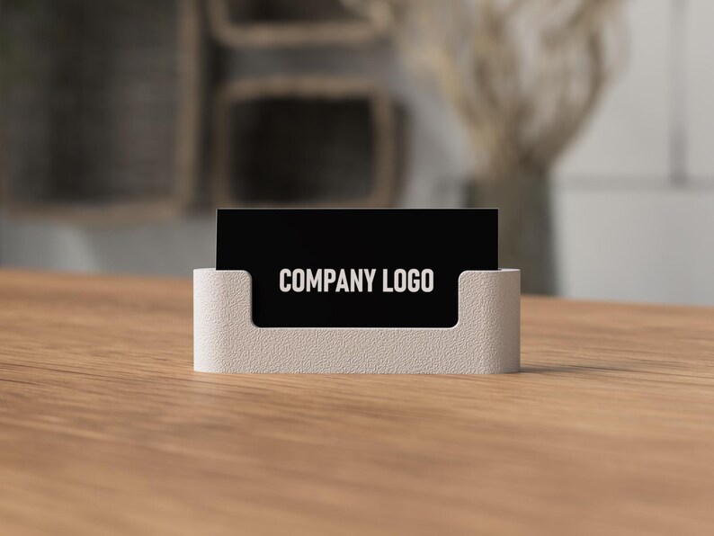 May include: A business card holder with a black card displaying "COMPANY LOGO" in white text. The holder is a light beige color and sits on a wooden surface. The background is blurred, suggesting an office or workspace setting.