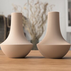 May include: Two beige vases with a modern design sit on a wooden surface. The vase on the left has a textured surface, while the one on the right has vertical ridges. Both have a wide base and a narrow neck.