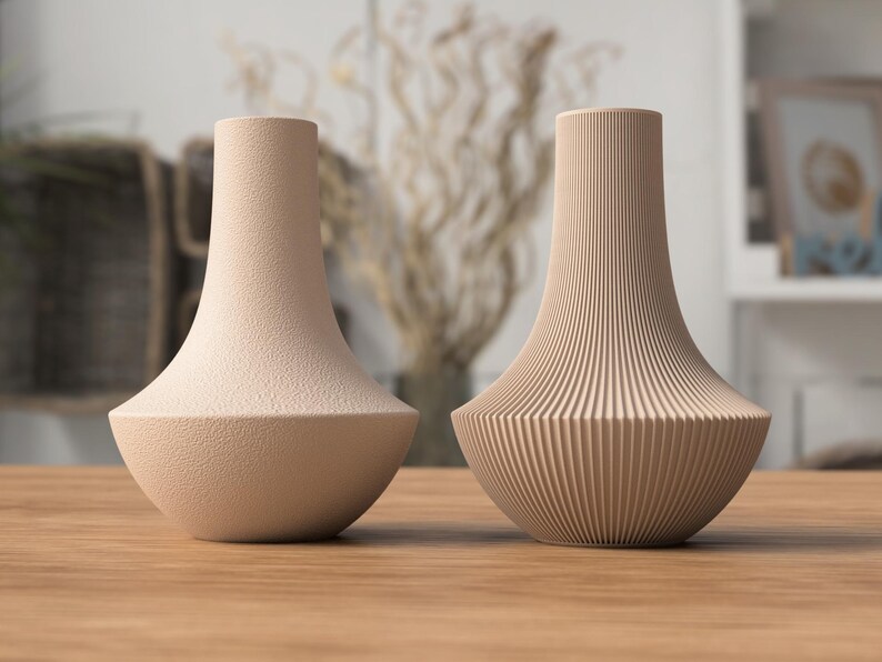 May include: Two beige vases with a modern design sit on a wooden surface. The vase on the left has a textured surface, while the one on the right has vertical ridges. Both have a wide base and a narrow neck.