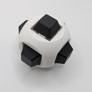 May include: A white and black puzzle toy. The toy is spherical with six black cube-shaped pieces protruding from a white, rounded frame. The toy is designed to be manipulated and solved.