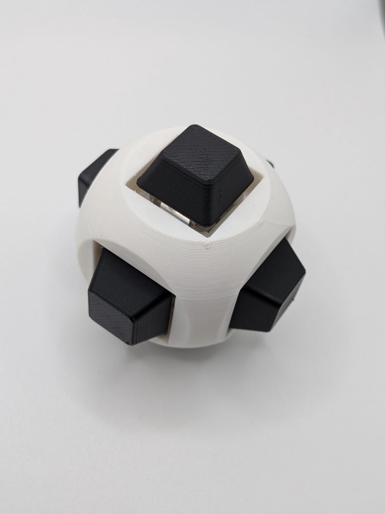 May include: A white and black puzzle toy. The toy is spherical with six black cube-shaped pieces protruding from a white, rounded frame. The toy is designed to be manipulated and solved.