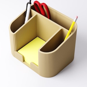 May include: A beige desk organizer with multiple compartments. It holds a yellow notepad, a white pen, red scissors, and a yellow pencil. The organizer has a ribbed texture and a curved design, suitable for office or home use.