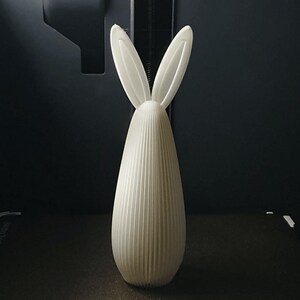 May include: A white, vertically-oriented decorative bunny figurine. The figurine has a long, oval body with vertical ridged detailing and two upright ears. The background is dark.