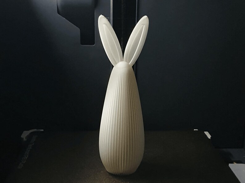 May include: A white, vertically-oriented decorative bunny figurine. The figurine has a long, oval body with vertical ridged detailing and two upright ears. The background is dark.