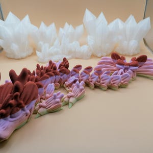 May include: A collection of 3D-printed fantasy sculptures. The image features a dragon with a gradient of pink, purple, and green, and white crystal formations. The dragon has a detailed head and body, with a textured surface. The crystals are sharp and angular.