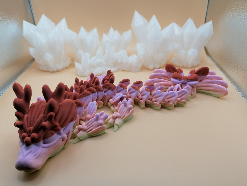 May include: A collection of 3D-printed fantasy sculptures. The image features a dragon with a gradient of pink, purple, and green, and white crystal formations. The dragon has a detailed head and body, with a textured surface. The crystals are sharp and angular.