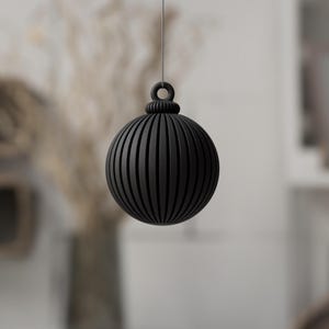 May include: A black, spherical ornament with vertical grooves, suspended by a thin string. The ornament has a decorative top with a loop for hanging. The background is blurred, suggesting a home decor setting.